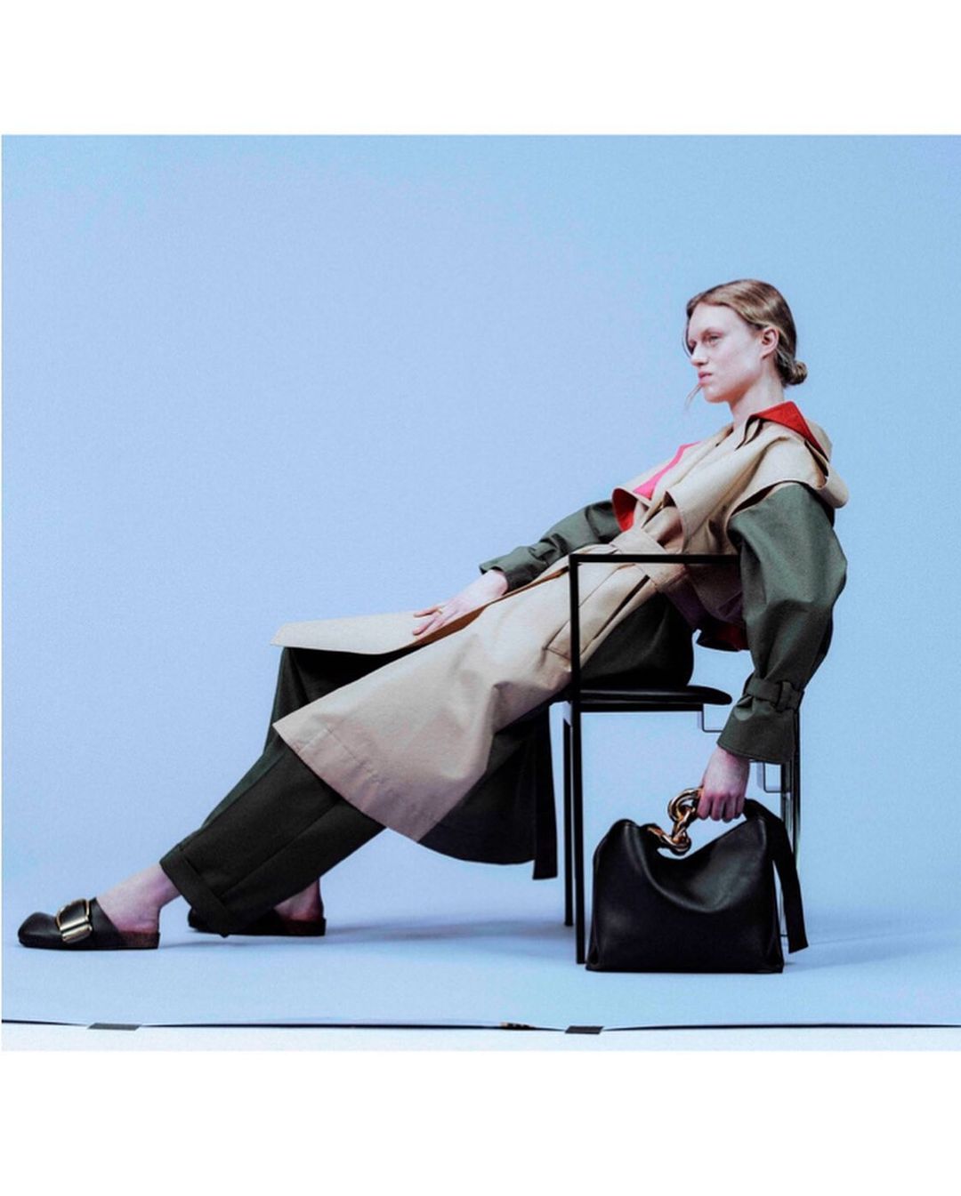 Woman in trench coat and dark pants seated on a black chair with a leather handbag against a gradient light blue background.
