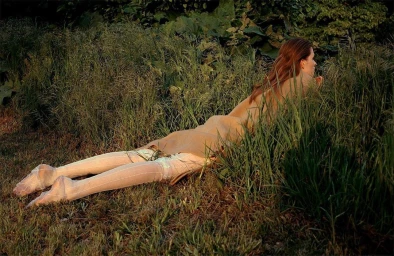 Woman with long red hair lying prone in tall green grass wearing sheer patterned tights and a tan top.