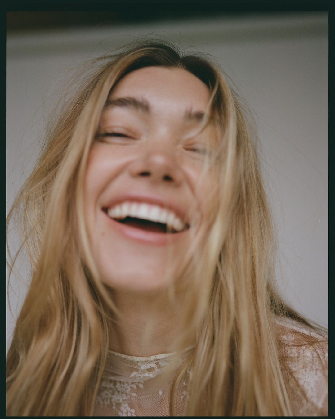 Close-up portrait of a smiling fair-skinned woman with long blonde hair, wearing a lace top against a neutral background. 