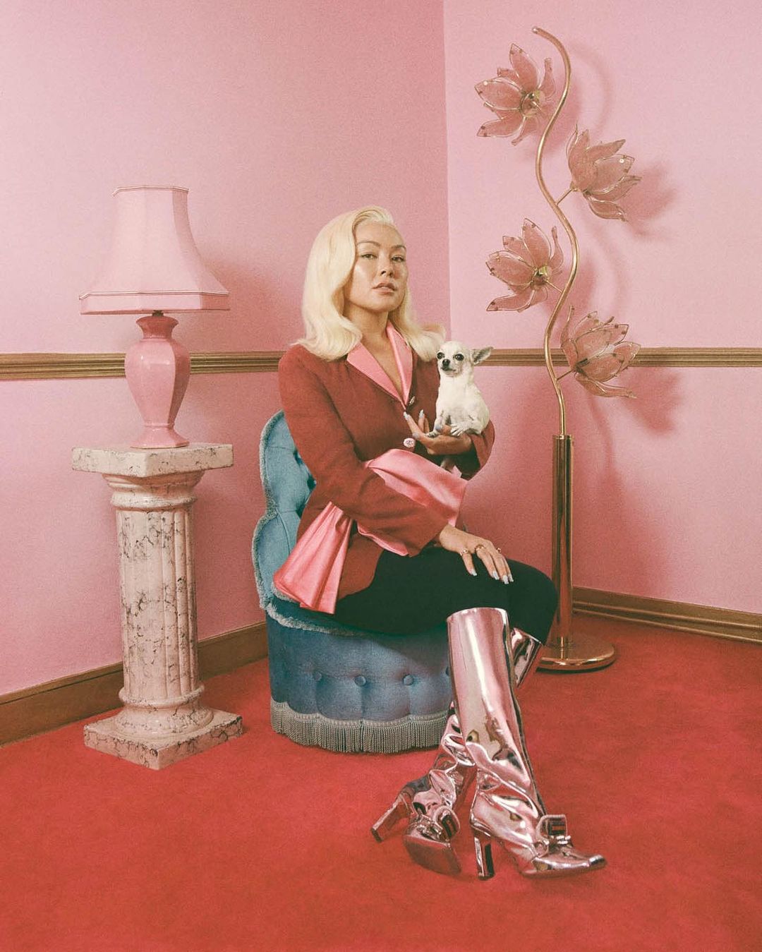Woman with platinum blonde hair in a pink interior, holding a chihuahua, seated next to a tall lamp and wearing shiny boots.