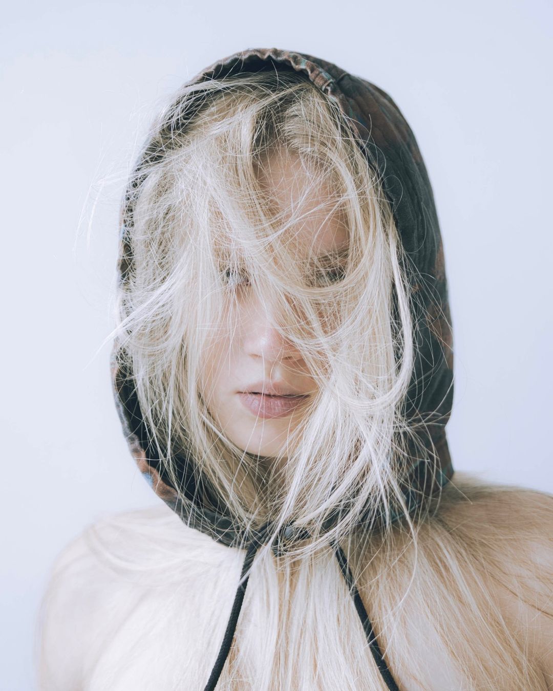 Close-up portrait of a woman with long blonde hair partially obscuring her face, wearing a hooded top against a neutral background.