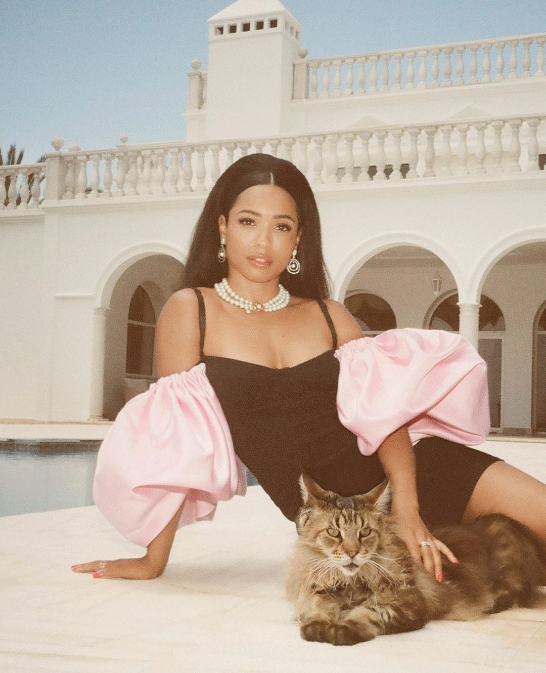Woman in black dress with pink sleeves sits with Maine Coon cat against a white building under clear sky in a glamorous pose.