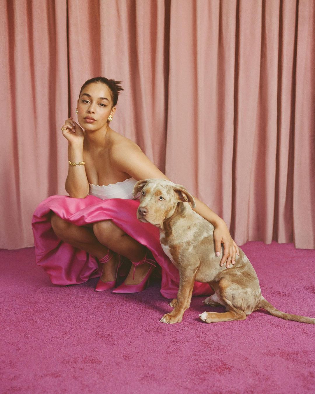A young woman and a dog are posing in a pink room with matching curtains and flooring, wearing a white top and pink skirt and shoes.