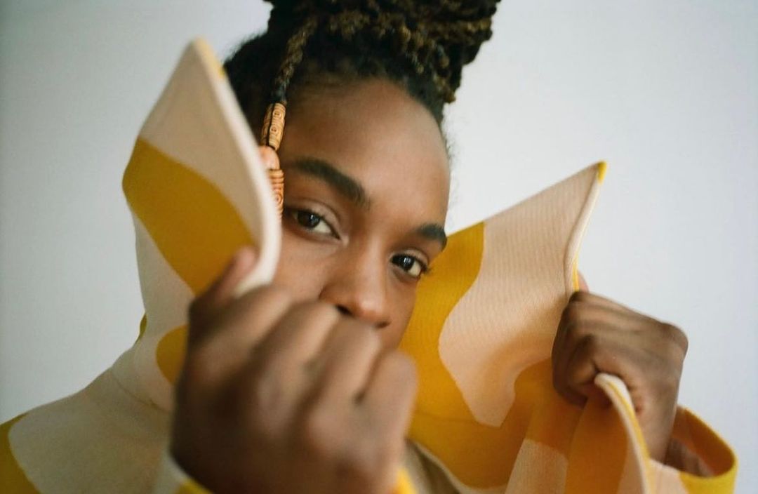 Close-up portrait of a Black woman with beaded braids, wearing a geometric yellow and cream-colored garment, looking directly at the camera.