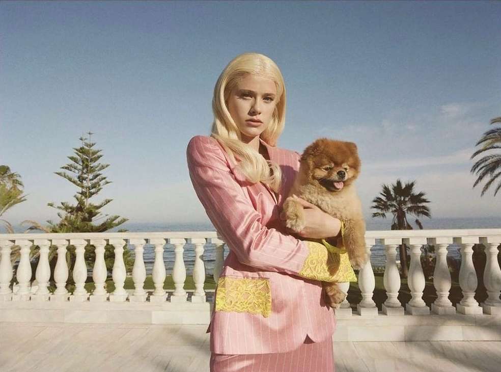 Woman in pink suit holds a Pomeranian dog outside on a sunny day. Palm trees and ocean are visible in the background.