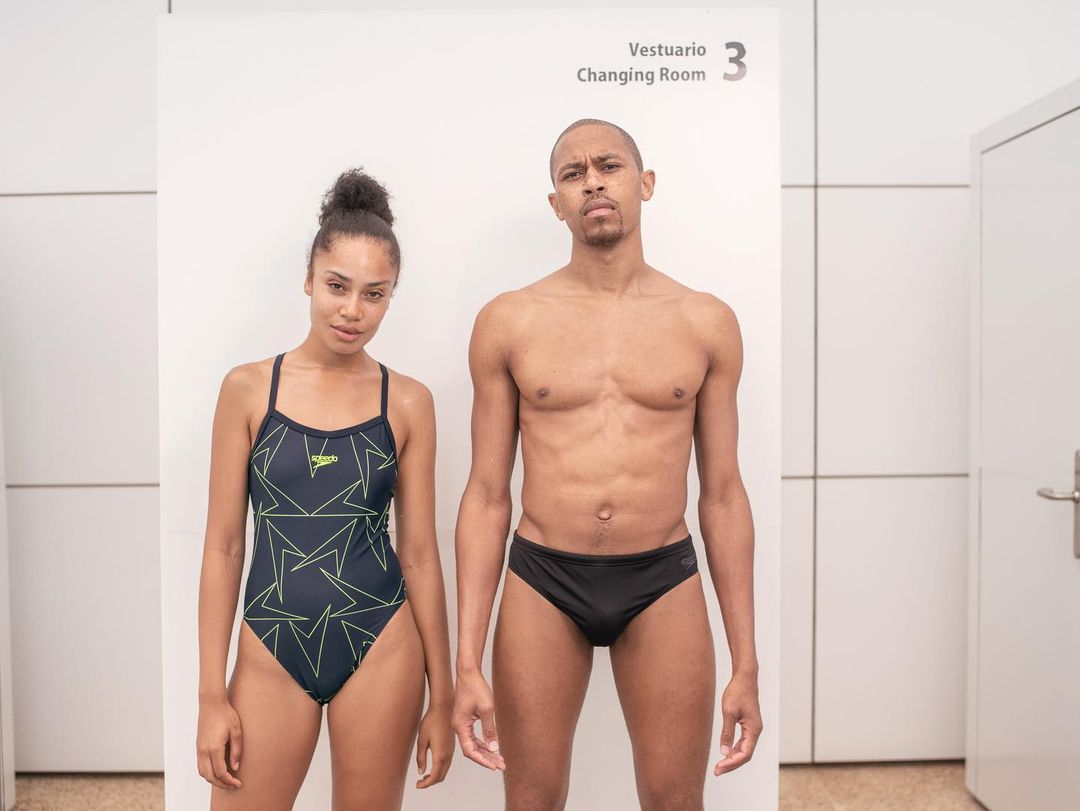 Two athletic adults in swimwear pose in a bright changing room. The woman wears a one-piece with patterns, while the man is shirtless.