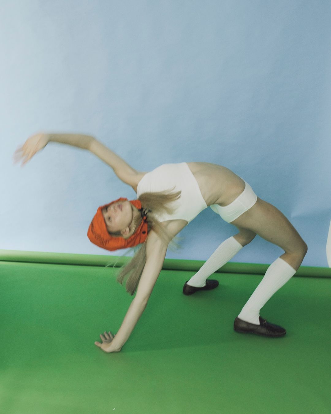 Adult woman in white bodysuit performs a backbend on a green surface in front of a light blue wall, captured in a studio setting.