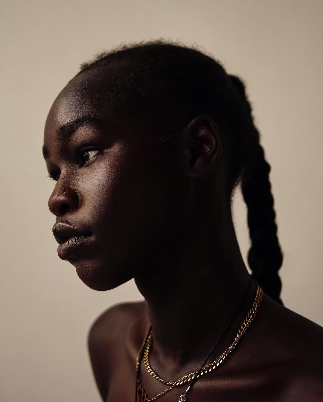 Portrait of a woman with dark skin in a profile view, showcasing layered gold necklaces and a simple, light-toned background.
