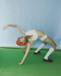Dancer in white crop top and shorts bending backward wearing an orange hat against blue and green studio backdrop