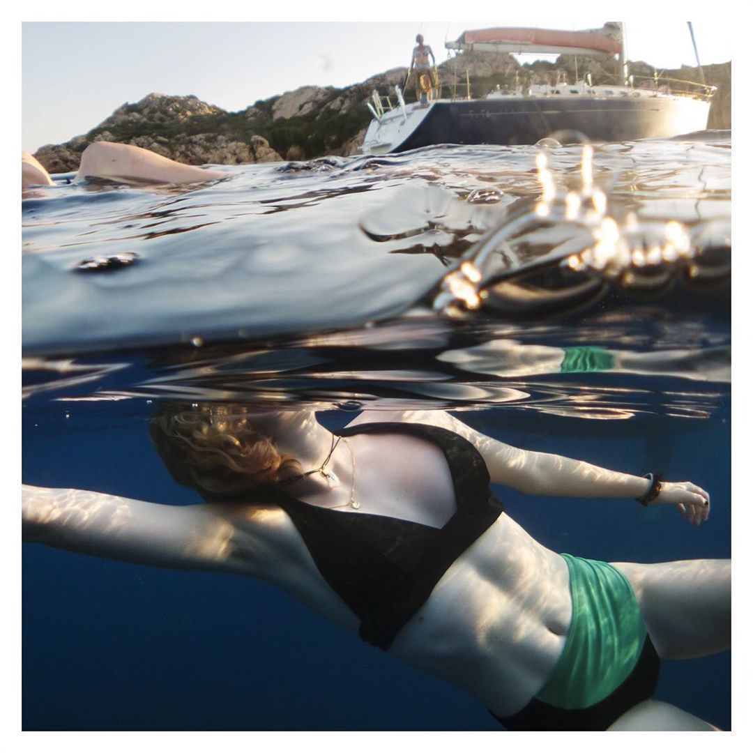 Underwater view of adult female swimming in a black bikini top and green shorts, with a boat and rocky shoreline in the background.