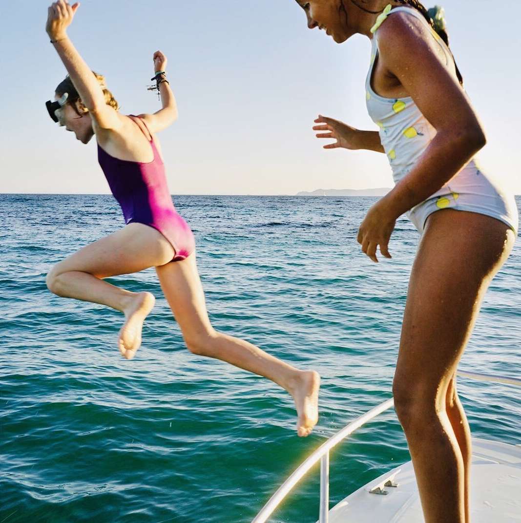 A young girl in a swimsuit jumps into the ocean from a boat, while another girl watches. Clear blue sky in the background.