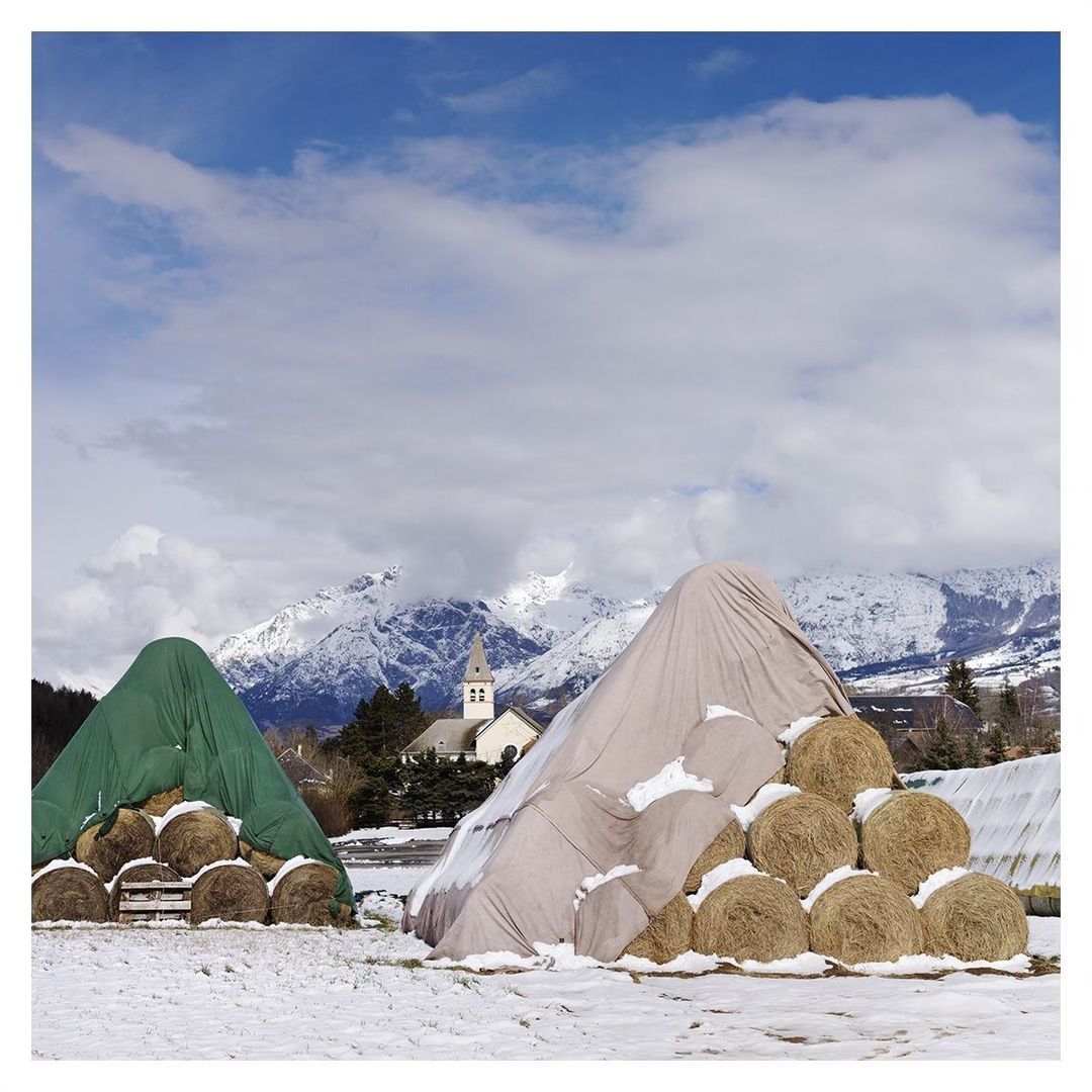 Winter scene of hay bales covered with tarps in a snow-covered field, with a church and mountains in the background under a cloudy sky.