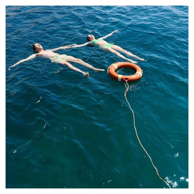 Two people in swimwear are floating on their backs in blue water next to a life preserver with a rope; water and blue sky background.
