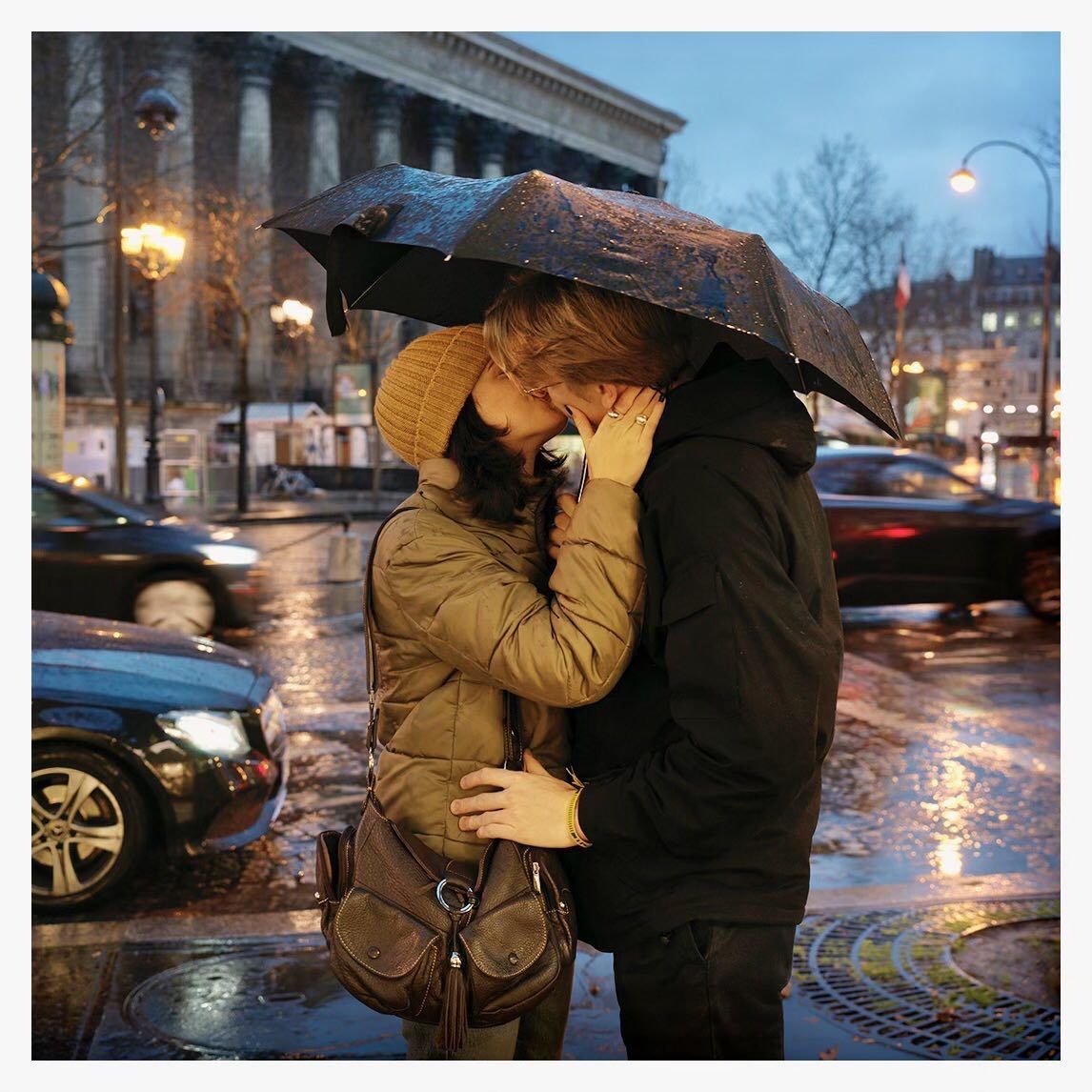 A couple kissing under an umbrella in the rain on a city street at night, with car traffic and building lights visible in the background.