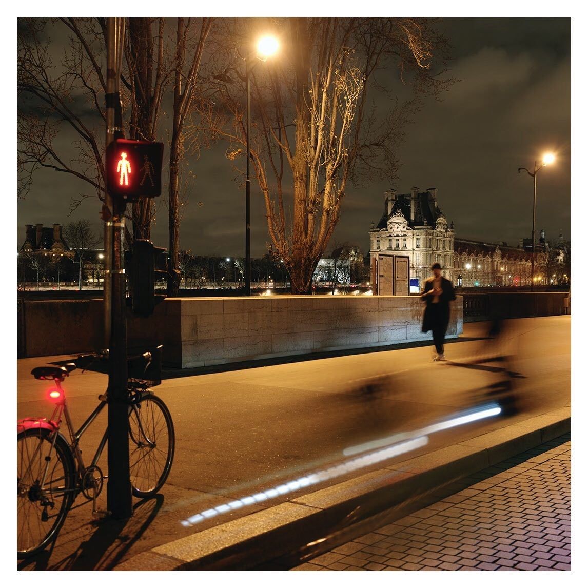 Night scene shows bicycle parked near pedestrian light, figure walking across the bridge, and distant building reflecting in river.