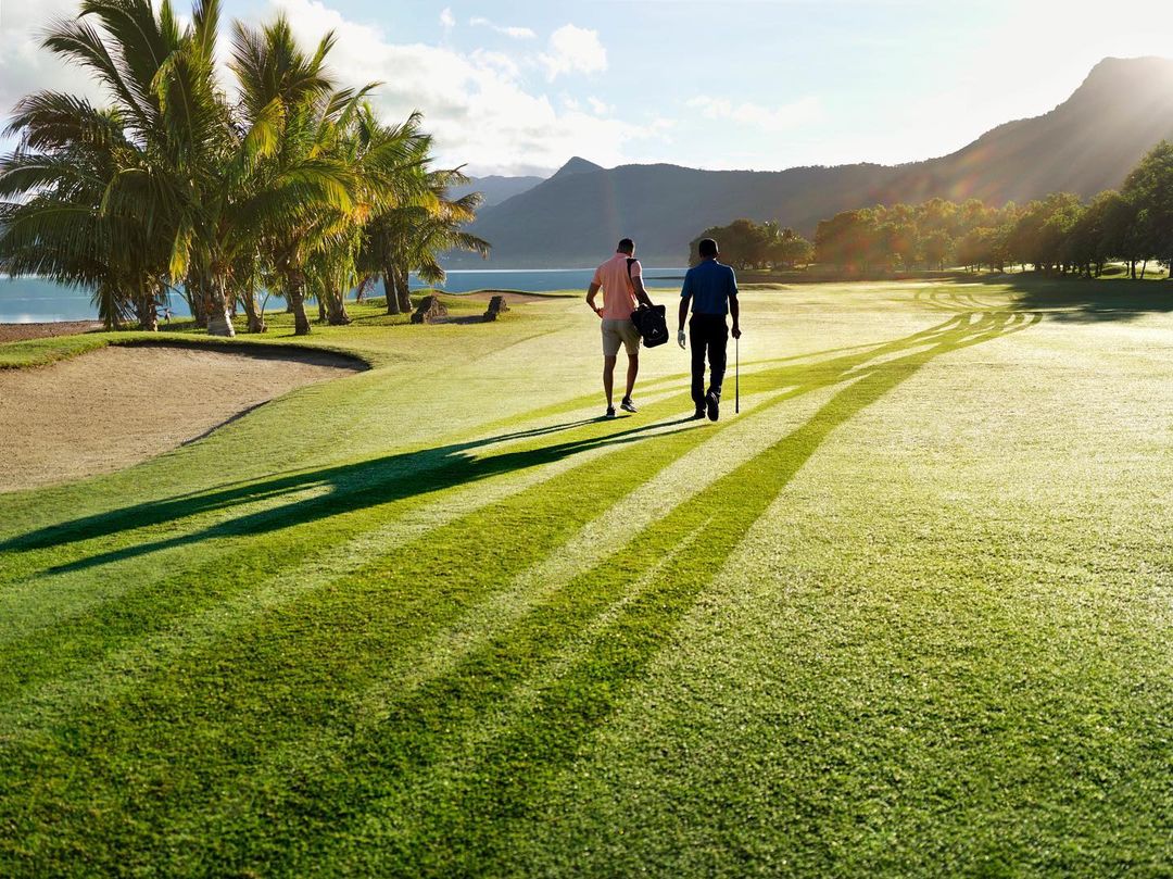 Two men walk on a golf course by the ocean, lined with palm trees, mountains in the background, bathed in soft morning light.
