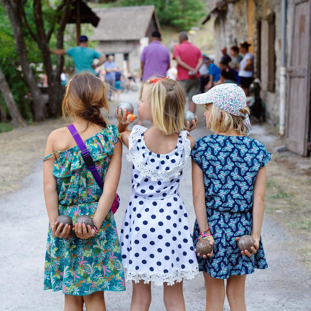 Three young girls stand outdoors, holding bocce balls, wearing dresses, and looking away in an outdoor setting with other people.