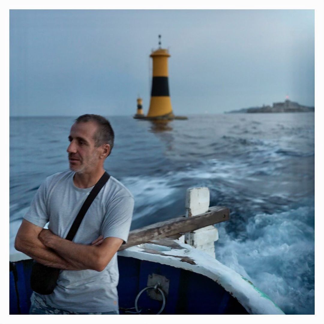 Man with crossed arms on a boat in the ocean with a yellow-and-black striped lighthouse and island in the background on a cloudy day.