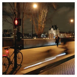 Bicycle parked next to a red pedestrian traffic signal at night with blurred motion of a cyclist on a city street.