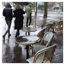 Wet bistro chairs and round tables on a rainy Paris street with pedestrians walking by under umbrellas.
