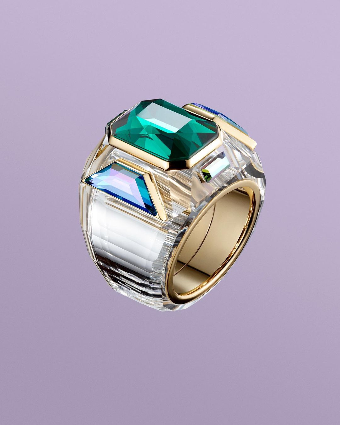 Chunky gold and clear crystal ring featuring a central emerald-cut green stone against a soft purple backdrop.