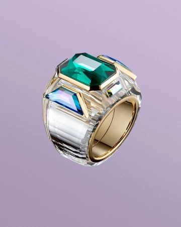 Chunky gold and clear crystal ring featuring a central emerald-cut green stone against a soft purple backdrop.