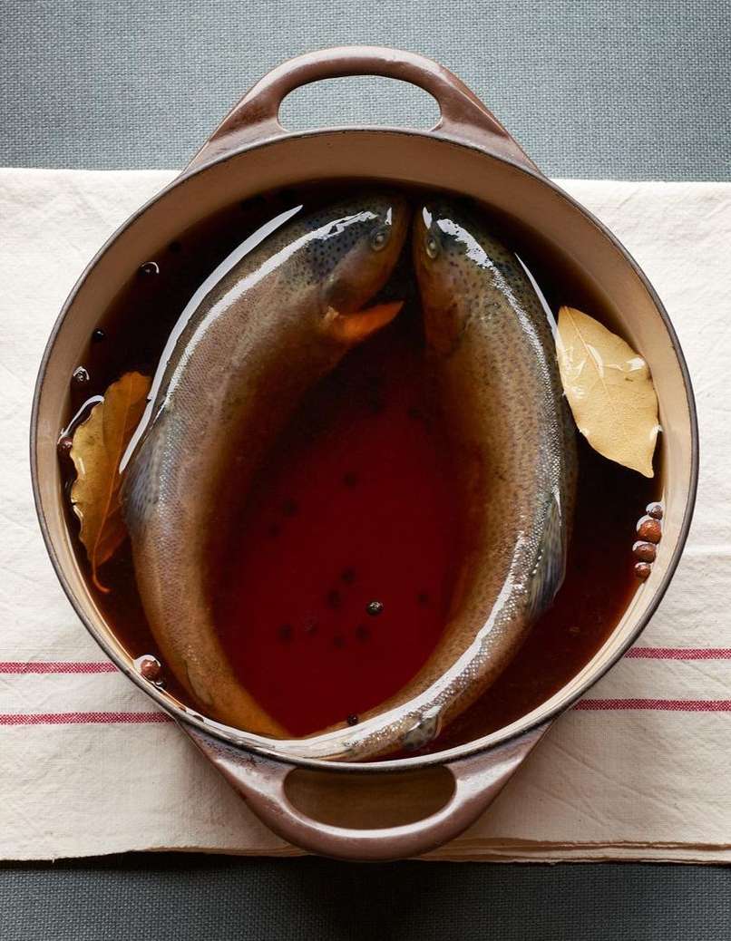 Overhead shot of two raw trout in a pot of dark liquid, seasoned with bay leaves and peppercorns, set on a white striped cloth.