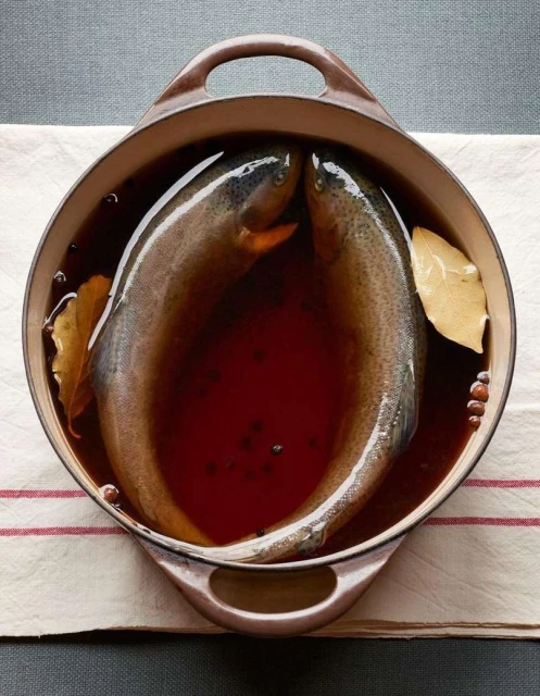 Overhead shot of two raw trout in a pot of dark liquid, seasoned with bay leaves and peppercorns, set on a white striped cloth.