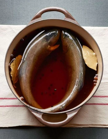 Overhead shot of two raw trout in a pot of dark liquid, seasoned with bay leaves and peppercorns, set on a white striped cloth.
