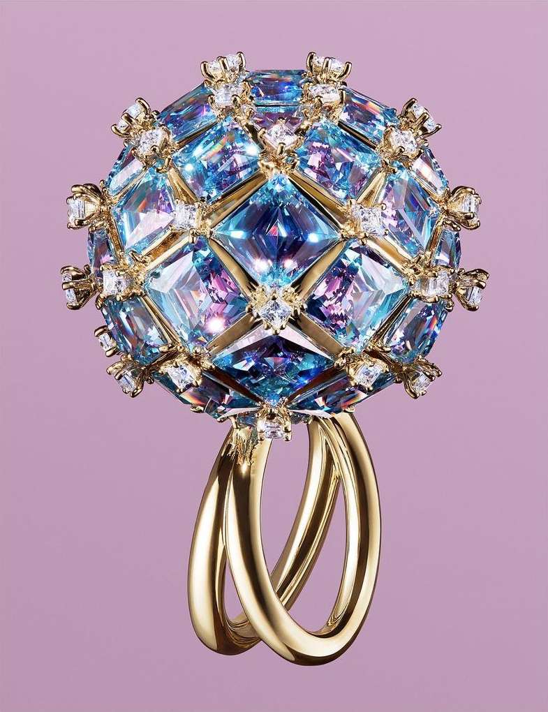 Close-up of a sphere-shaped ring featuring gold bands and encrusted with blue gems and small white diamonds against a pale purple background.