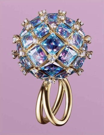 Close-up of a sphere-shaped ring featuring gold bands and encrusted with blue gems and small white diamonds against a pale purple background.