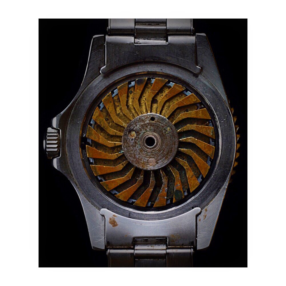 Close-up of a stainless steel wrist watch with a gold-colored, fan-shaped internal mechanism, set against a stark black background.