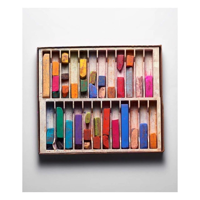 Overhead shot of a box filled with an array of colorful art pastels arranged neatly in rows against a white background.