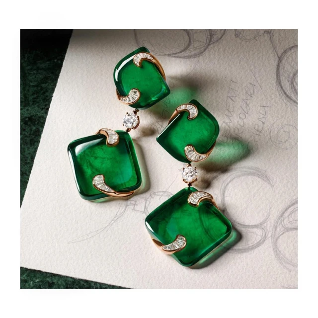 Elegant studio shot of emerald-green, square-cut drop earrings with diamond accents on a cream-colored surface featuring faint pencil sketches.