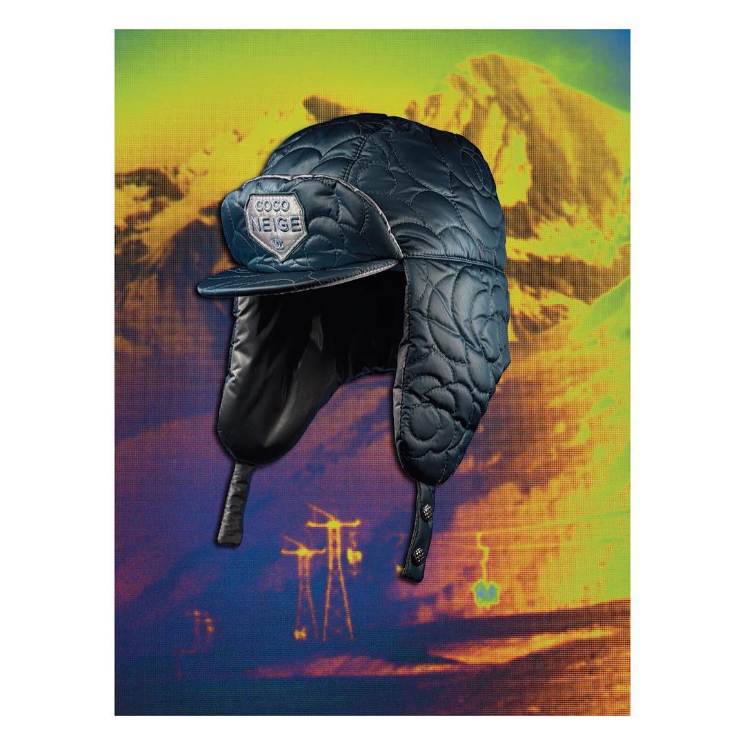Close-up of a blue quilted trapper hat with ear flaps and a logo against a vibrant yellow and blue mountain backdrop.