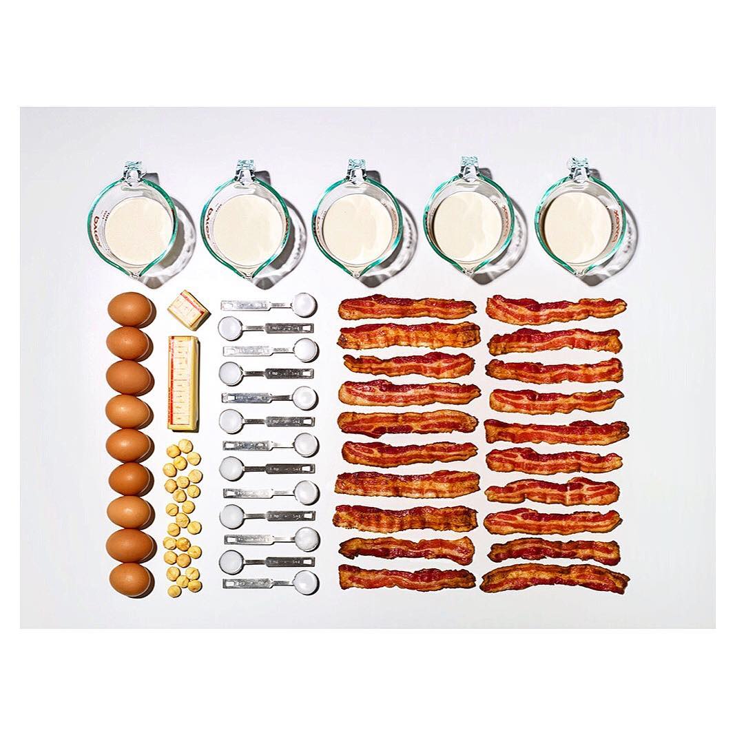 Overhead shot of baking ingredients arranged on a white surface: eggs, milk, butter, bacon strips, and measuring spoons.