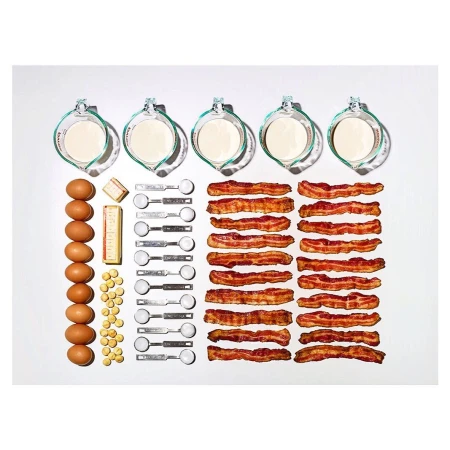 Overhead shot of baking ingredients arranged on a white surface: eggs, milk, butter, bacon strips, and measuring spoons.
