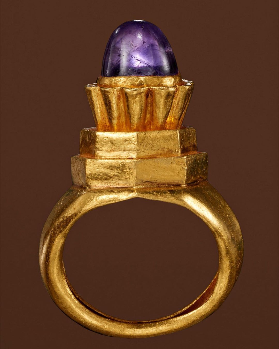 Gold ring with a purple cabochon gemstone, studio shot against a brown backdrop, jewellery design, close up view.