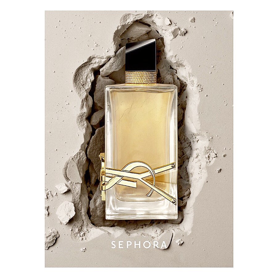 A rectangular clear glass Yves Saint Laurent perfume bottle filled with amber liquid sits amidst a bed of light beige powder and shards.