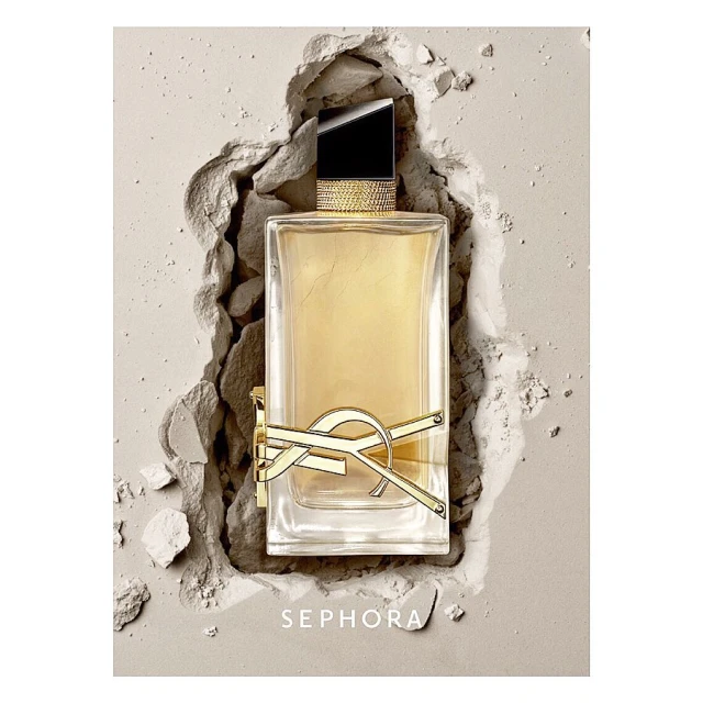 A rectangular clear glass Yves Saint Laurent perfume bottle filled with amber liquid sits amidst a bed of light beige powder and shards.