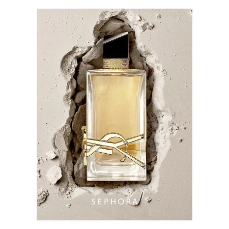 A rectangular clear glass Yves Saint Laurent perfume bottle filled with amber liquid sits amidst a bed of light beige powder and shards.