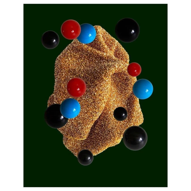Abstract still life featuring a textured, golden, folded form surrounded by floating blue, red, and black spheres against a dark green backdrop.