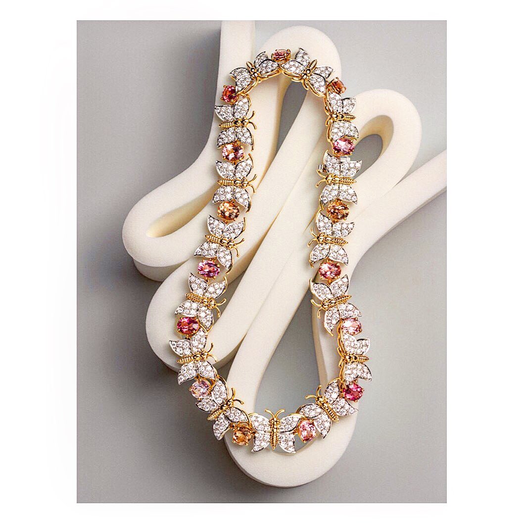 Elegant gold necklace featuring butterfly and flower motifs adorned with sparkling white and pink gemstones, displayed on a soft gray backdrop.