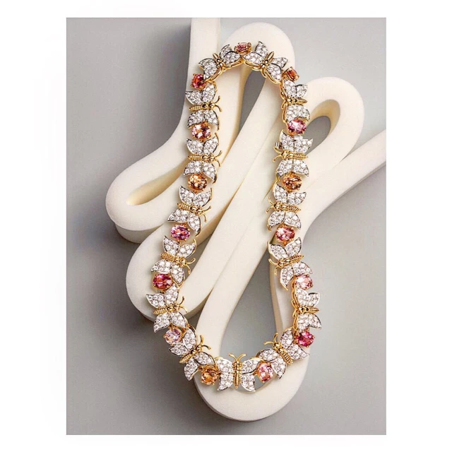 Elegant gold necklace featuring butterfly and flower motifs adorned with sparkling white and pink gemstones, displayed on a soft gray backdrop.