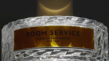 Close-up of a crystal perfume bottle with a gold label reading 'Room Service Vilhelm Parfumerie New York' on a dark background.