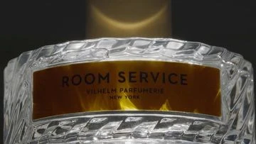 Close-up of a crystal perfume bottle with a gold label reading 'Room Service Vilhelm Parfumerie New York' on a dark background.