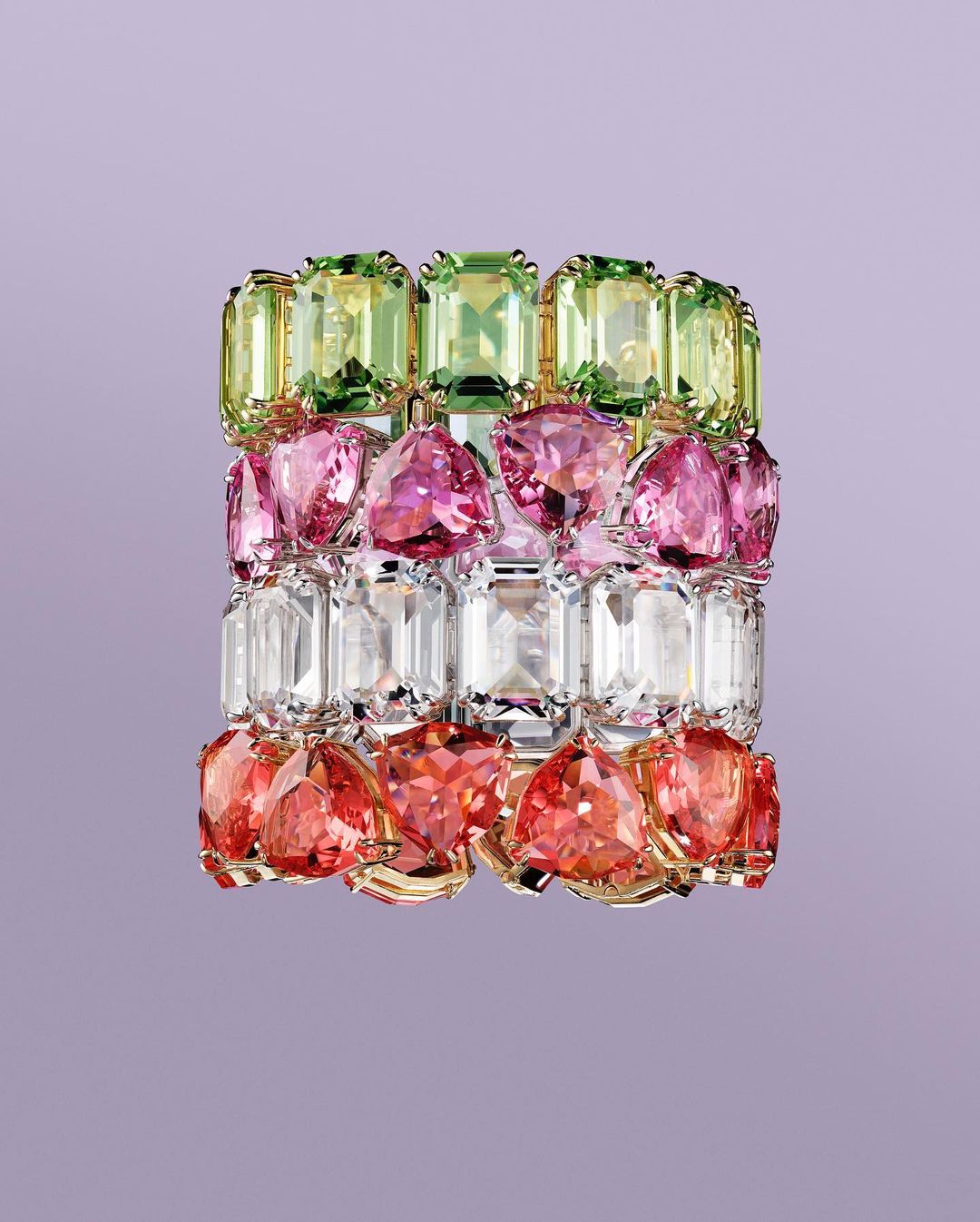 Colorful gemstone bracelet featuring green, pink, clear, and red stones set in gold, against a light purple backdrop. Jewelry still life.