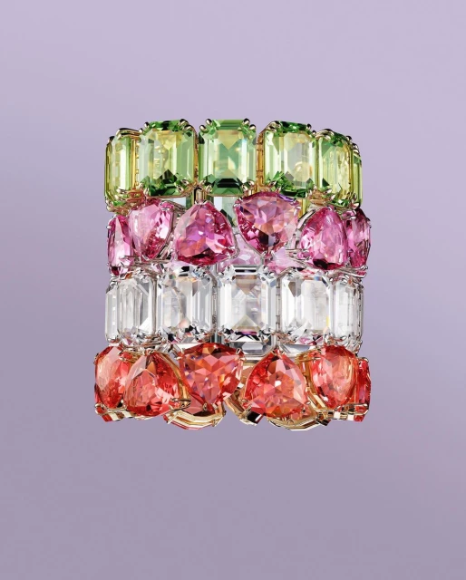 Colorful gemstone bracelet featuring green, pink, clear, and red stones set in gold, against a light purple backdrop. Jewelry still life.