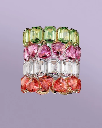 Colorful gemstone bracelet featuring green, pink, clear, and red stones set in gold, against a light purple backdrop. Jewelry still life.
