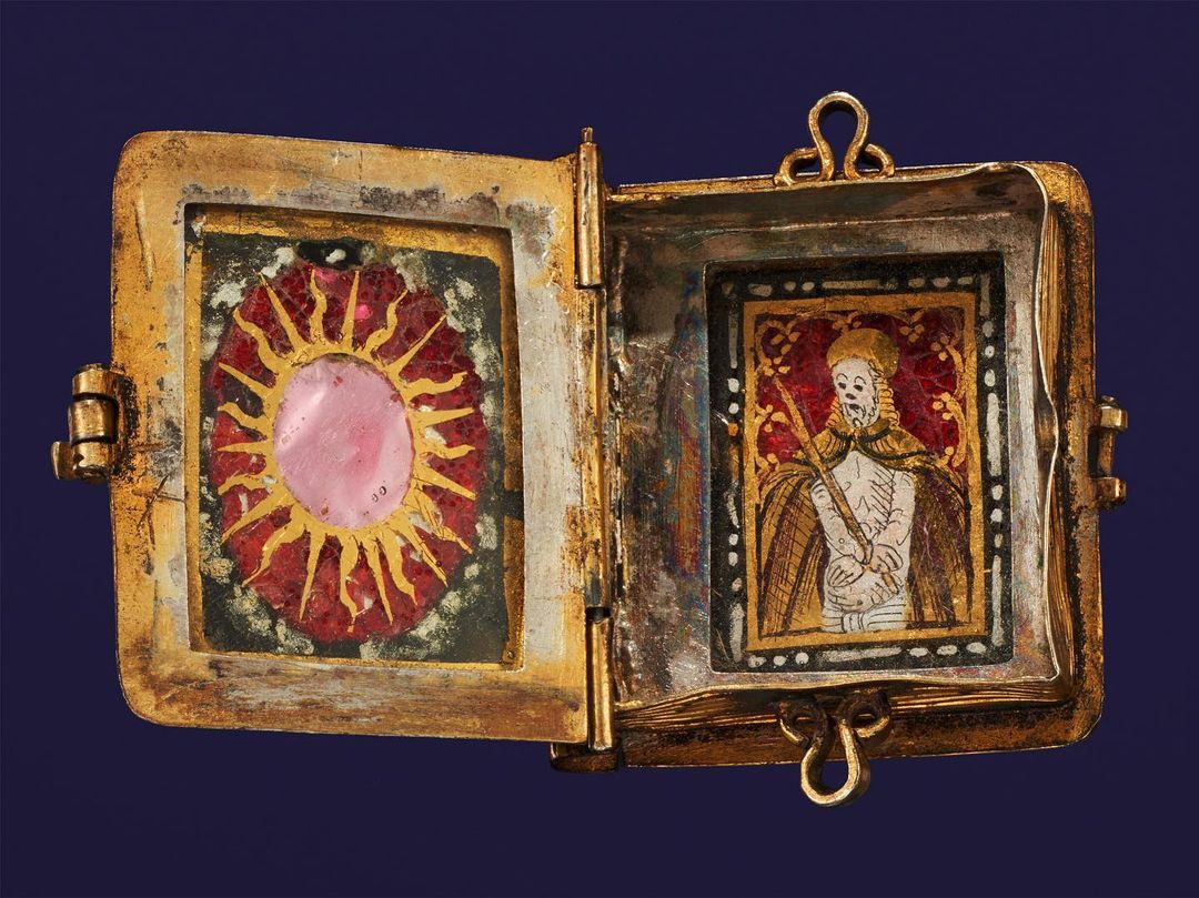 Open golden locket showcasing religious art, one side featuring a pink sun-like image, the other an illustrated religious figure.